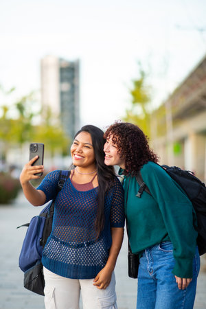 Two young women standing outdoors, smiling as they take a selfie with a smartphone. Both are wearing casual clothes and carrying backpacks, suggesting they are on a trip or an outing in the cityの写真素材
