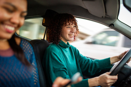 Woman smiling while driving a car with a female friend in the passenger seat. The driver is focused on the road, and the passenger is out of focus, smiling and holding a phoneの写真素材