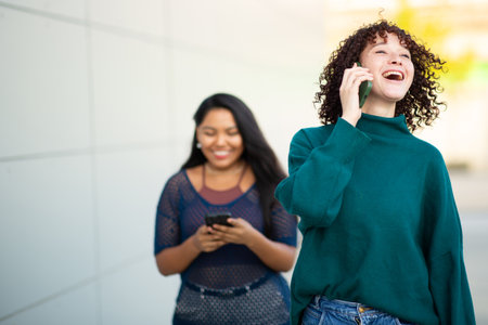 A young woman with curly hair laughs while talking on the phone as she walks outdoors, followed by another woman who is focused on texting on her smartphoneの写真素材