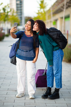 Two young women, one taking a selfie with a smartphone, standing on a city street with backpacks and luggage. They are dressed casually, suggesting they are traveling or on an urban adventureの写真素材