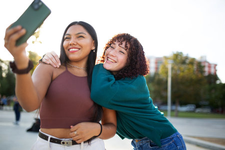 Two women standing outdoors in a park, smiling as they take a selfie with a smartphone. One woman holds the phone while the other hugs her from behind, with trees and sunlight in the backgroundの写真素材