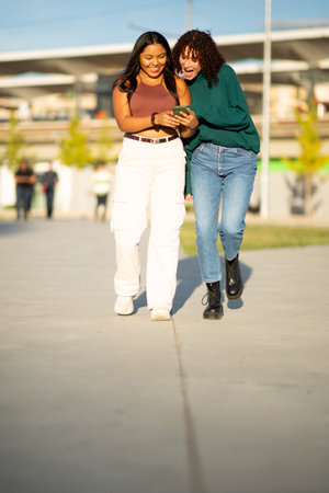 Two young women walking side by side on a sunny day, looking at a phone and laughingの写真素材