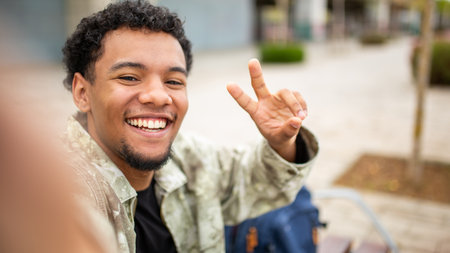 Young man with curly hair smiling and showing a peace sign gesture with his fingers while taking a selfie outsideの写真素材