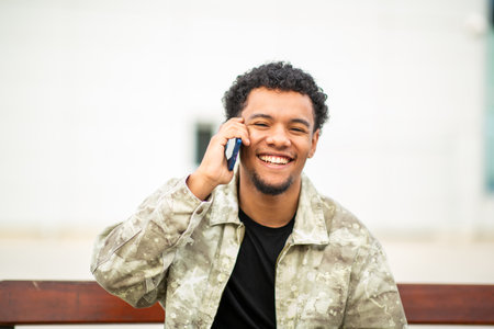 Portrait of a young man sitting outdoors on a bench, smiling while talking on his smartphoneの写真素材