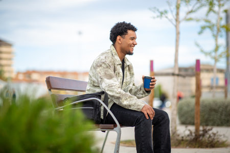 Man seated on a bench holding a blue coffee cup, with a black bag beside him, enjoying the outdoors in an urban setting including trees and buildingsの写真素材