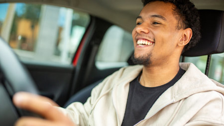 Young man smiling as he drives a car, holding the steering wheel with a blurred cityscape visible through the windowの写真素材