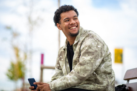 Young man sitting outdoors, smiling while holding a smartphone in his hand and looking backの写真素材