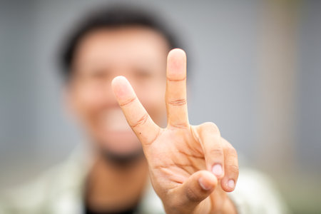 Close-up of a hand showing a peace sign in focus, with the young man's smiling face blurred in the backgroundの写真素材
