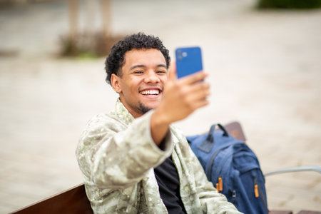 Young man sitting on a bench in an urban area, smiling widely while holding his smartphone for a selfieの写真素材