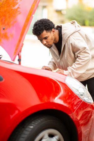 Young man bending over to examine the engine of a red car with the hood open, focused on identifying the mechanical issueの写真素材