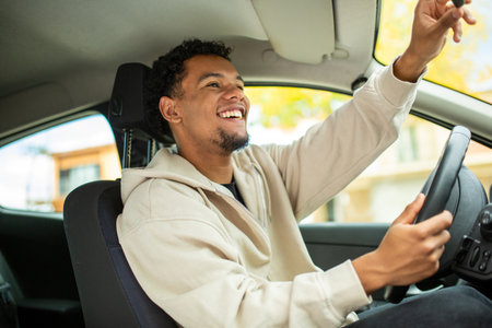 Man in beige hoodie smiling and adjusting the rear-view mirror inside a car, with one hand on the steering wheelの写真素材