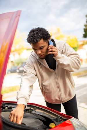 Young man examining the engine of a red car with the hood open while talking on his mobile phoneの写真素材
