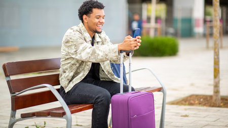 Side view of a smiling young man sitting on a bench with a purple suitcase, holding a smartphoneの写真素材
