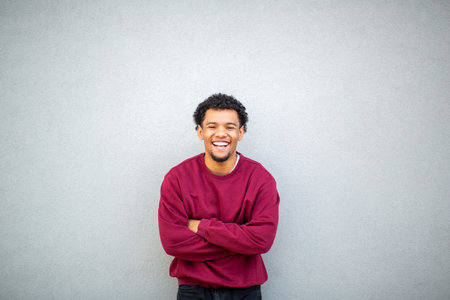 Young man wearing a red sweater, smiling with arms crossed while standing against a gray wallの写真素材