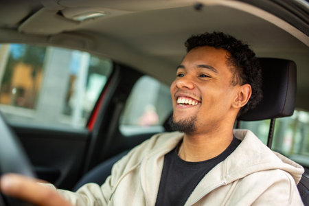 Young man in a beige hoodie smiling and looking forward while driving a car, with hand on the steering wheelの写真素材