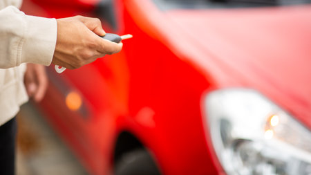 Close-up of a hand holding a car key in front of a red car, focusing on the key with the car's headlight and red body blurred in the backgroundの写真素材