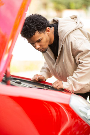 Young man in a beige hoodie leaning forward to inspect the engine of a red car with the hood openの写真素材