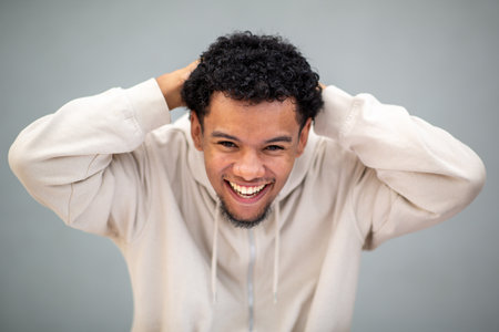 Young man wearing a beige hoodie, smiling broadly with hands resting on his head, standing against a plain gray backgroundの写真素材