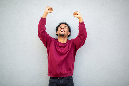 Young man wearing a red sweater, smiling with raised fists in a celebratory pose against a gray wallの写真素材