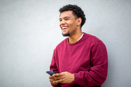 Young man with curly hair and a cheerful expression holding his smartphone in a casual poseの写真素材