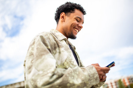 Smiling young man holding his smartphone while standing in a bright, open area with a blue sky in the backgroundの写真素材