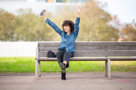 A young curly-haired woman in a blue shirt and black jeans sitting on a wooden bench in a park, smiling brightly with arms raised in excitement while holding a smartphoneの写真素材