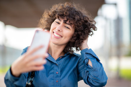 Portrait of a curly-haired woman wearing a blue shirt, smiling while touching her hair as she takes...
