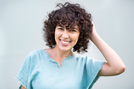 Close-up of a young woman with curly hair smiling warmly while touching her hair and wearing a light...