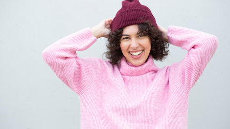 Vibrant portrait of a cheerful young woman wearing a bright pink sweater and a burgundy knit beanie, posing against a light wall and smiling warmlyの写真素材