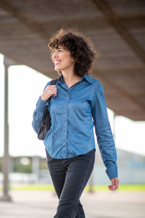 Portrait of a young woman with curly hair, smiling while walking outdoors in a blue shirt and black...