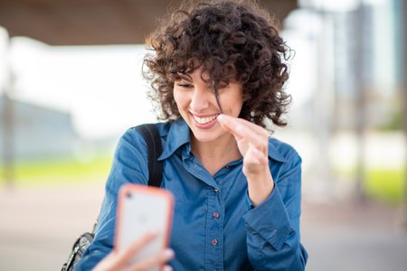 Woman with curly hair and blue shirt smiling while playing with her hair and holding a phone during...