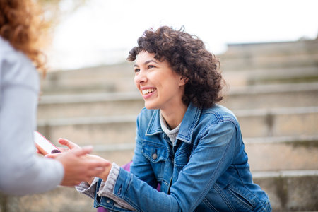 Young woman with curly hair wearing a denim jacket smiling while talking to someone holding a...