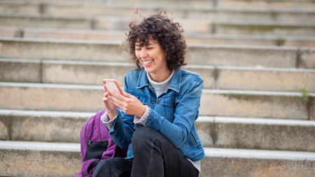Young woman with curly hair wearing a denim jacket and sweater, sitting on stone steps with a purple...