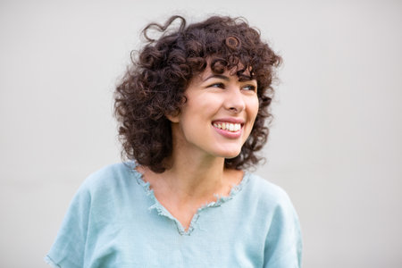 Close-up of a young woman with curly hair smiling and looking away while wearing a light blue top against a plain white backgroundの写真素材