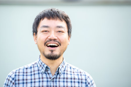 Close-up portrait of an Asian man smiling with open mouth in a checkered shirt against a plain...
