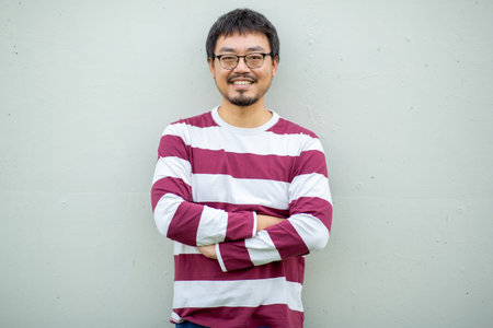 Portrait of a mature man with a beard, wearing glasses and a striped t-shirt standing with folded...