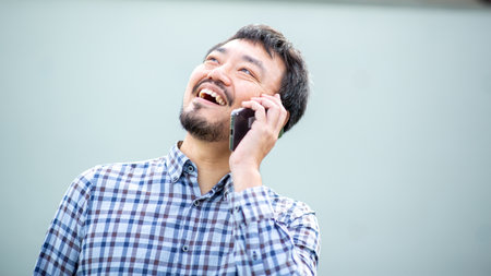 Portrait of a smiling Asian man wearing a checkered shirt while looking upwards and talking on his...