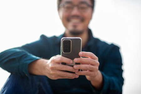 Close-up view of hands holding a smartphone with a transparent case, with a blurred smiling man in glasses and teal shirt in the background, captured in an outdoor settingの写真素材