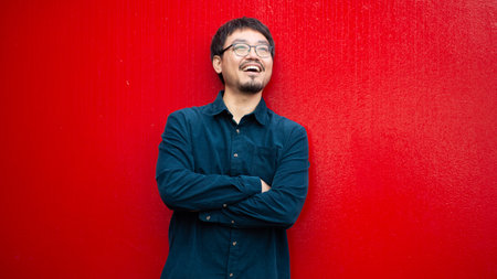 Portrait of an Asian man wearing glasses and a dark shirt, smiling with arms crossed while standing...