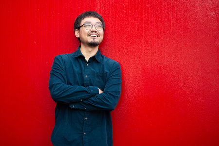 Asian man wearing glasses and a dark shirt, smiling confidently while standing against a vibrant red wallの写真素材