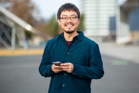 Portrait of a Taiwanese man wearing glasses and a dark teal shirt smiling while holding a smartphone in both hands, standing outdoors with blurred urban setting in the backgroundの写真素材