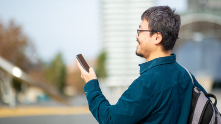 Side view of an Asian man wearing glasses, a teal shirt, and a backpack, holding a phone while...