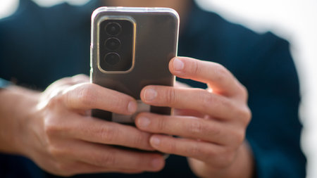 Close-up view of hands holding a smartphone with a transparent case, showing the camera module, in an outdoor setting with blurred backgroundの写真素材