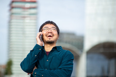 Portrait of a man with glasses and a teal shirt smiling broadly while holding a phone to his ear, with blurred modern urban area in the backgroundの写真素材
