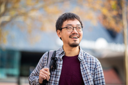 A cheerful Asian man wearing glasses and a checkered shirt with a backpack, looking away and smiling...