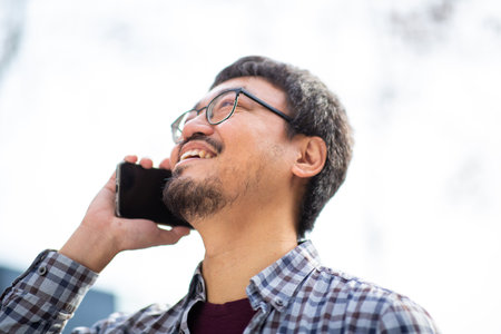 A close-up side view of an Asian man wearing glasses and a plaid shirt, smiling while talking on a...