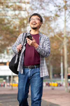 A Taiwanese man in a plaid shirt and maroon t-shirt holding a phone and smiling while walking outdoors with a backpack. The background features an urban park with blurred trees and pavementの写真素材