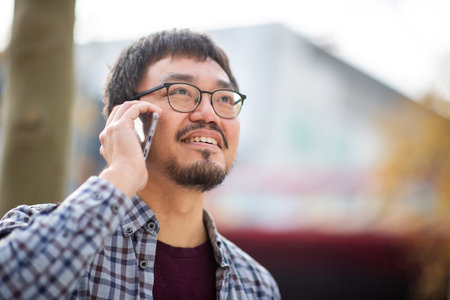 Portrait of an Asian man wearing glasses and a plaid shirt talking on a phone while standing outdoors with blurred backgroundの写真素材