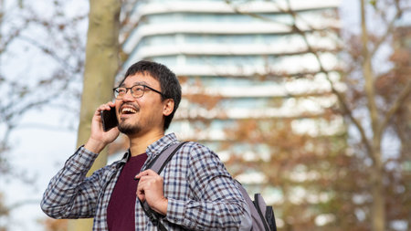 An Asian man wearing glasses and casual clothing talking on a phone and smiling while holding a...