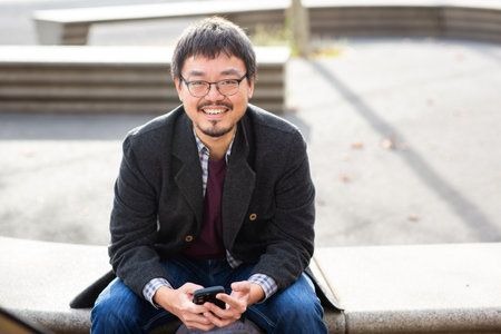 Smiling mature Asian man wearing glasses and a gray coat sitting on a bench outdoors, holding a...